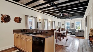 Een moderne keuken met een vaatwasser, houten kasten en een groot eiland. Er is een zithoek met een tafel en stoelen, en een woonkamer met een bank en een plafondventilator.