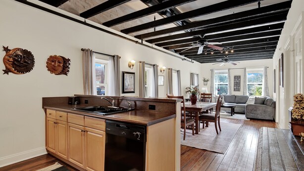 A modern kitchen with a dishwasher, wooden cabinets, and a large island. There is a dining area with a table and chairs, and a living room with a sofa and a ceiling fan. - Stay Next Door to City Market, Heated Pool Access (Savannah)