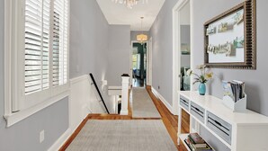 A hallway with a chandelier, a white console table with magazines, and a framed photo collage on the wall. - Walkable Home in Thomas Square, Heated Pool Access (Savannah)