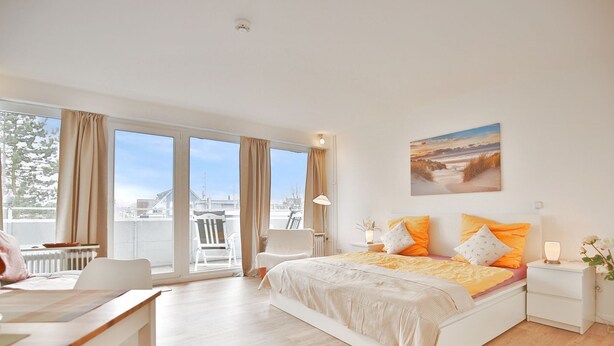 Une chambre avec un grand lit, un bureau et une chaise, une vue sur la ville grâce à de grandes fenêtres, et un tableau accroché au mur.