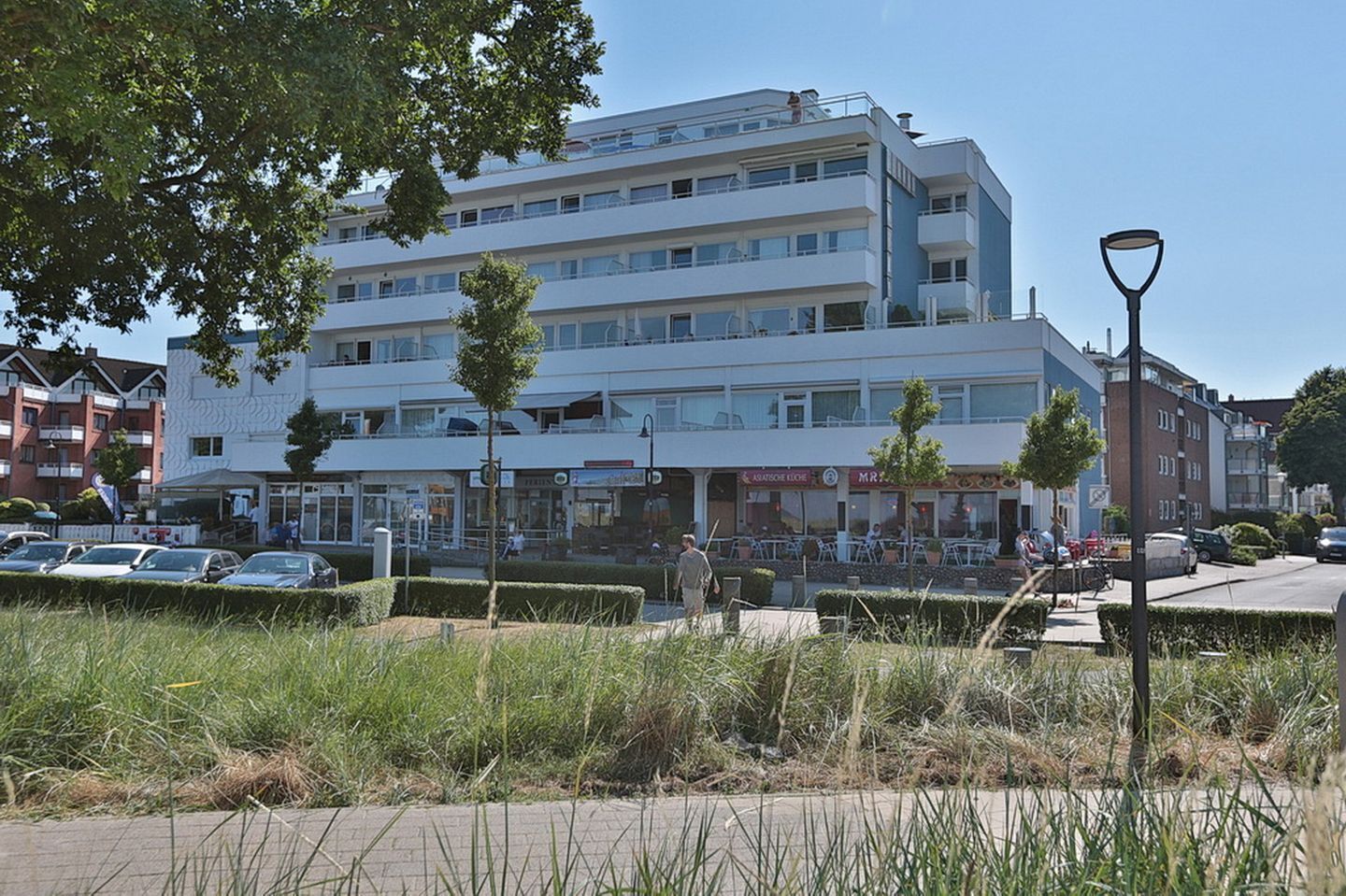 A multi-story building with a flat facade, several ground-level shops, and a parking area with cars.