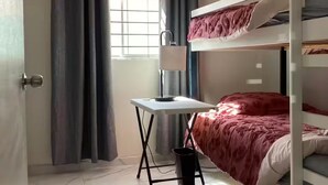 Una habitación con una cama individual alta, un escritorio con lámpara, un cesto de basura y una ventana con cortinas.
