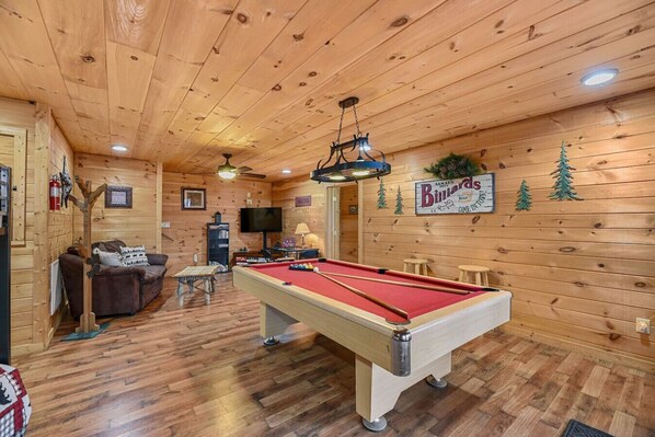 Un intérieur en bois comprenant une table de billard, un canapé, un téléviseur et un panneau indiquant « Billiards ».
