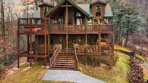 A two-story wooden cabin with a green roof, surrounded by trees and a wooden deck. - Big Blue Ridge | MTN Views, Hot Tub, Pool Table! (Sevierville)