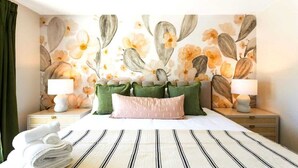 A hotel room with a bed, pillows, a nightstand, a lamp, and a wall with a floral pattern.