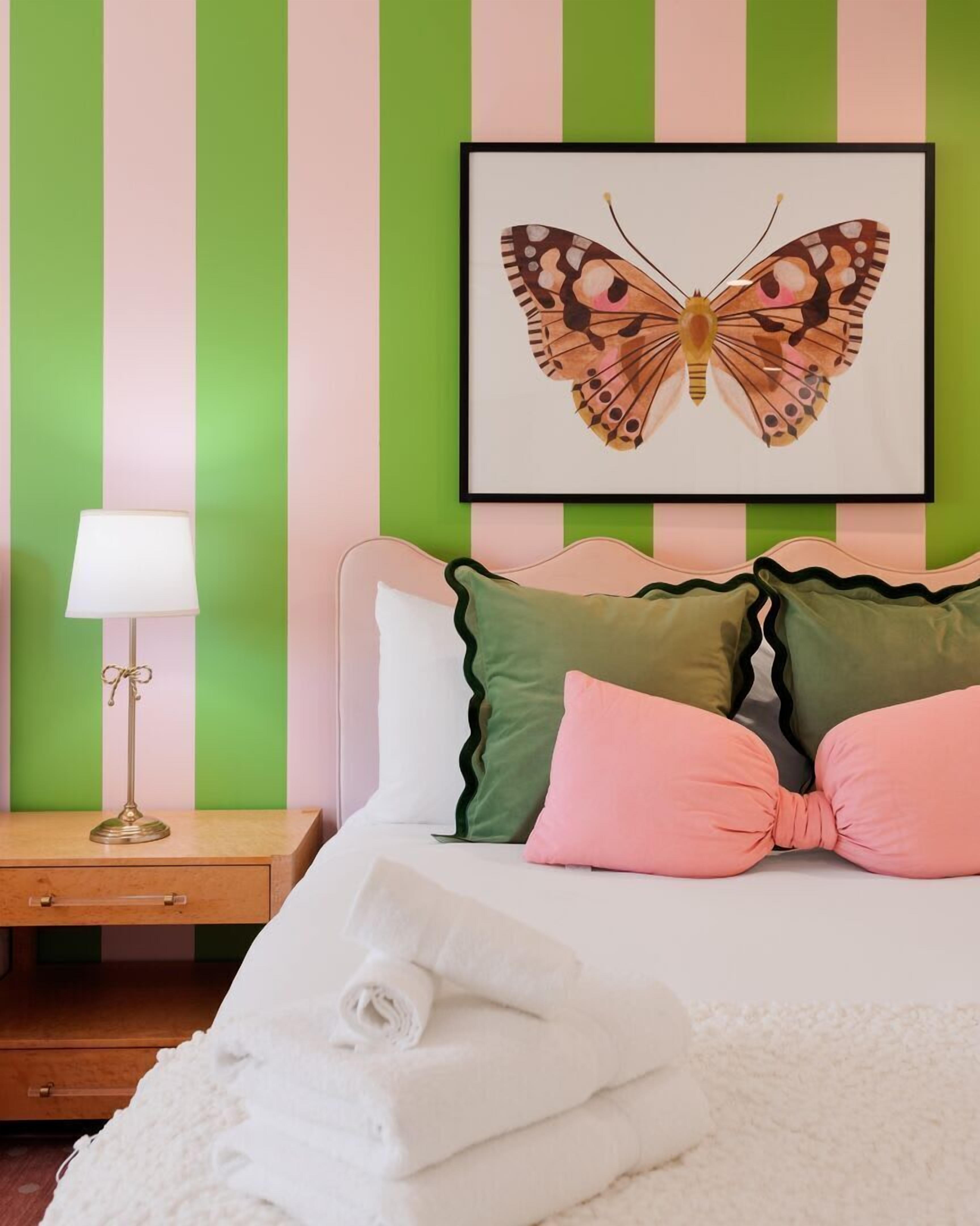 A bedroom with striped walls, a framed butterfly artwork, a bedside lamp, a nightstand, a drawer, and a bed with pillows.