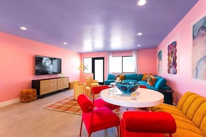 Una sala de estar con una mesa redonda, sillas rojas, un sofá azul y un televisor en la pared.