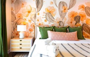 Un dormitorio con papel tapiz floral, una mesita de noche con lámpara y una cama con ropa de cama y almohadas a rayas.