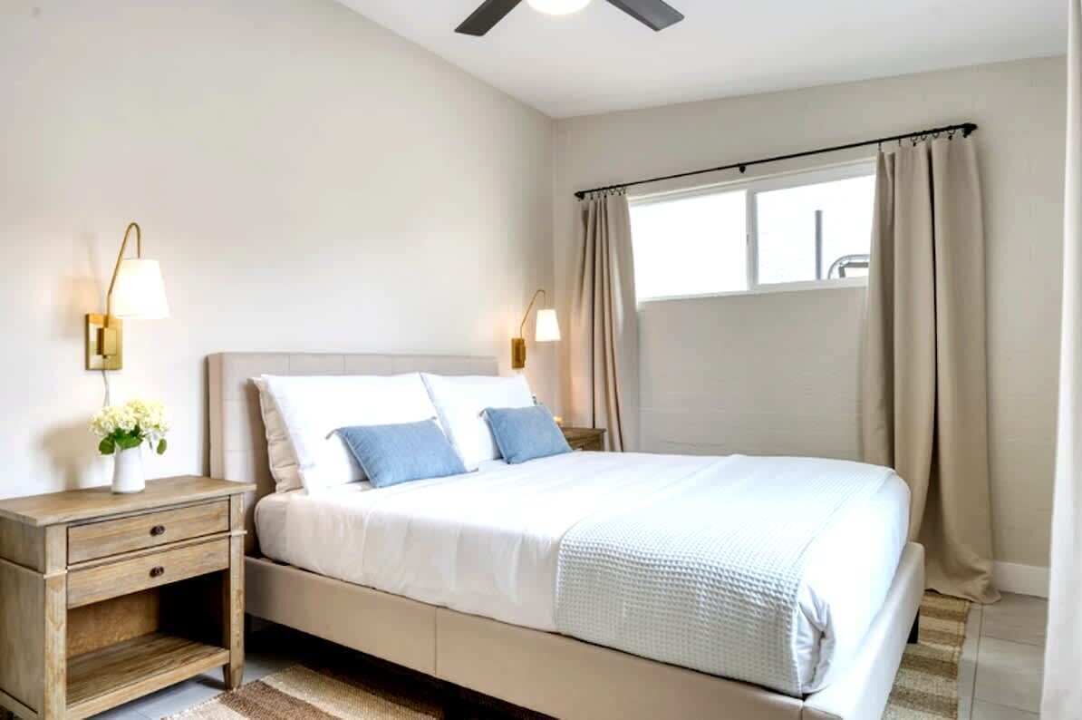 Ein Schlafzimmer mit Bett, Nachttischen, Lampen, einem Fenster mit Vorhängen und einem Deckenventilator.