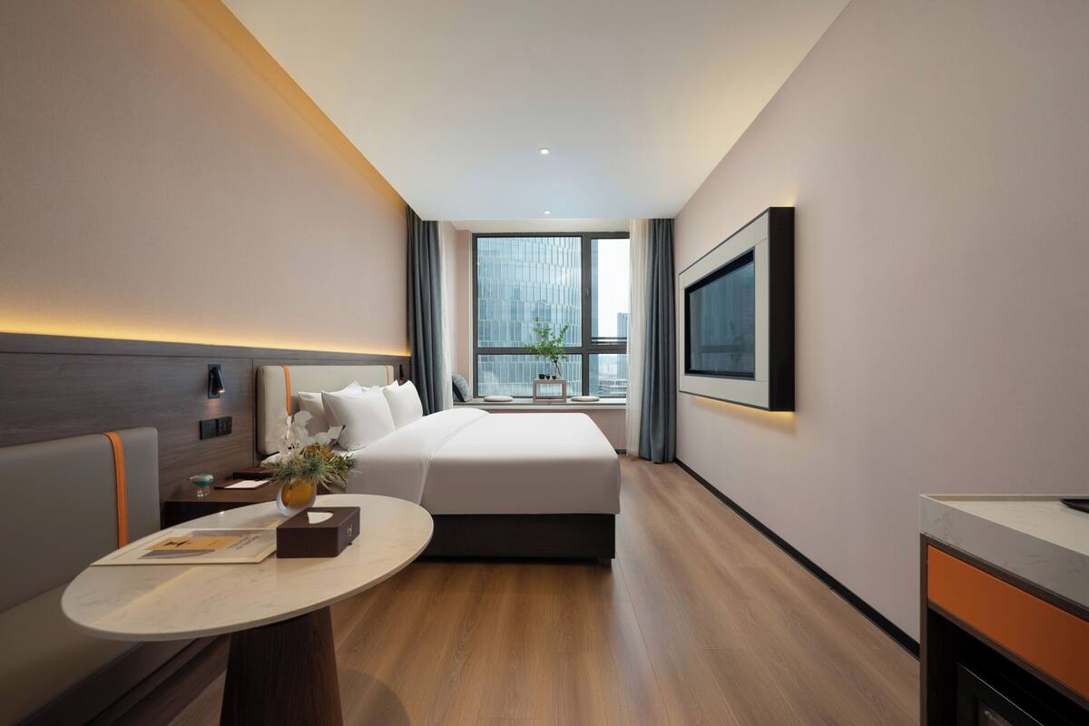 Ein modernes Hotelzimmer mit einem großen Bett, einem Schreibtisch, einem Fernseher und einem Fenster mit Blick auf die Stadt.