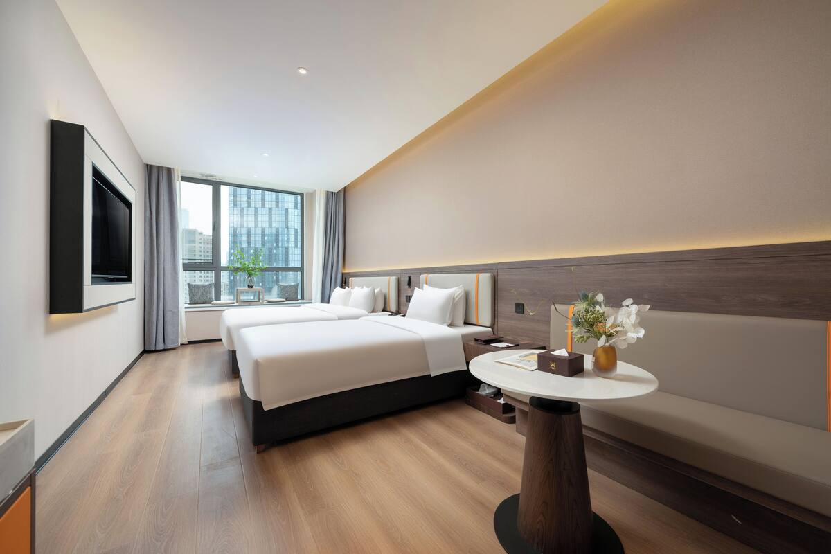 Ein modernes Hotelzimmer mit einem großen Bett, einem an der Wand montierten Fernseher, einem runden Tisch mit Blumenvase und einem großen Fenster mit Blick auf die Stadt.