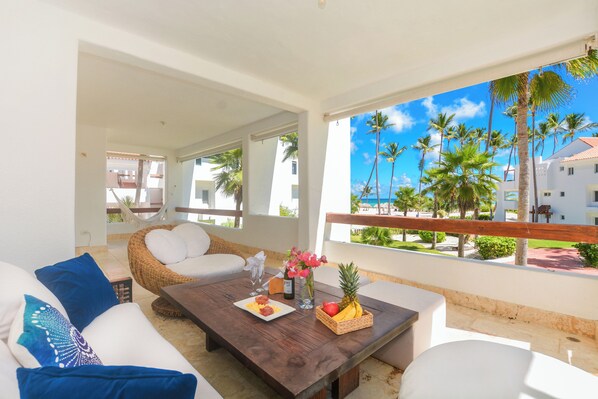 A balcony with a wooden table, white cushions, and a wicker chair.