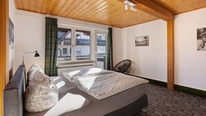 A bedroom with a wooden ceiling, a bed with floral bedding, a window with curtains, a chair, and a picture on the wall.