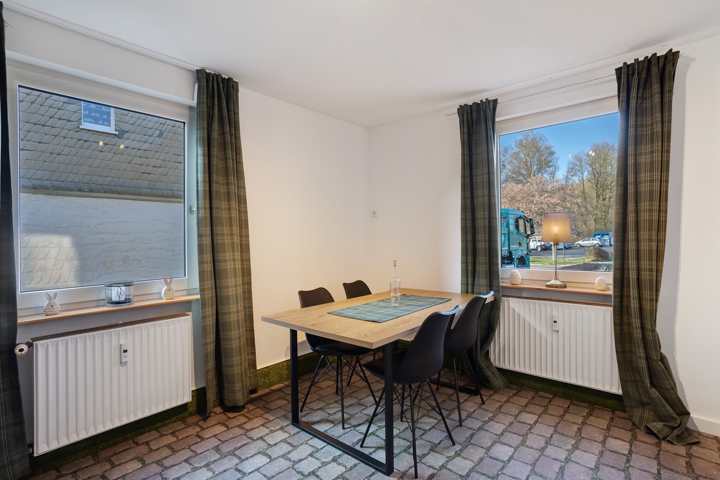 Ein Zimmer mit Tisch und Stühlen, einem fenster mit Vorhängen, einem Heizkörper und Blick ins Freie.