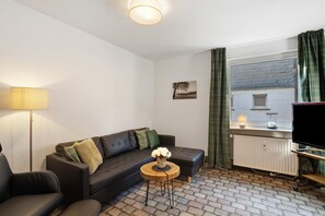 A living room with a sofa, a small round table with a vase, a lamp, a radiator, a TV, and a window with curtains.