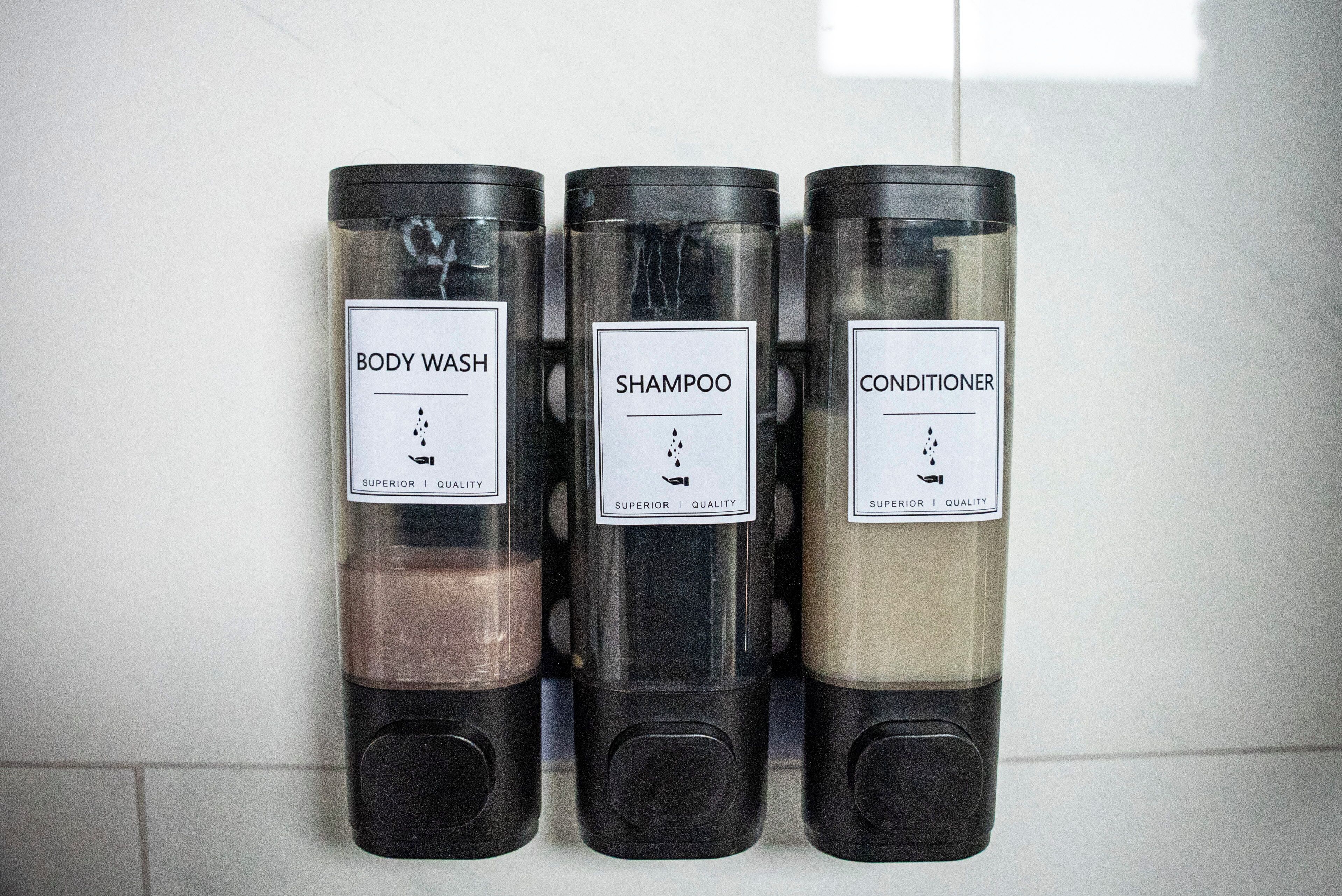 Three black dispensers labeled BODY WASH, SHAMPOO, and CONDITIONER, each with a white label and a clear section showing the product inside.