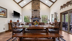 Un vaste salon comprenant une grande table à manger en bois, des fauteuils en cuir, une cheminée en pierre et un téléviseur fixé au mur.