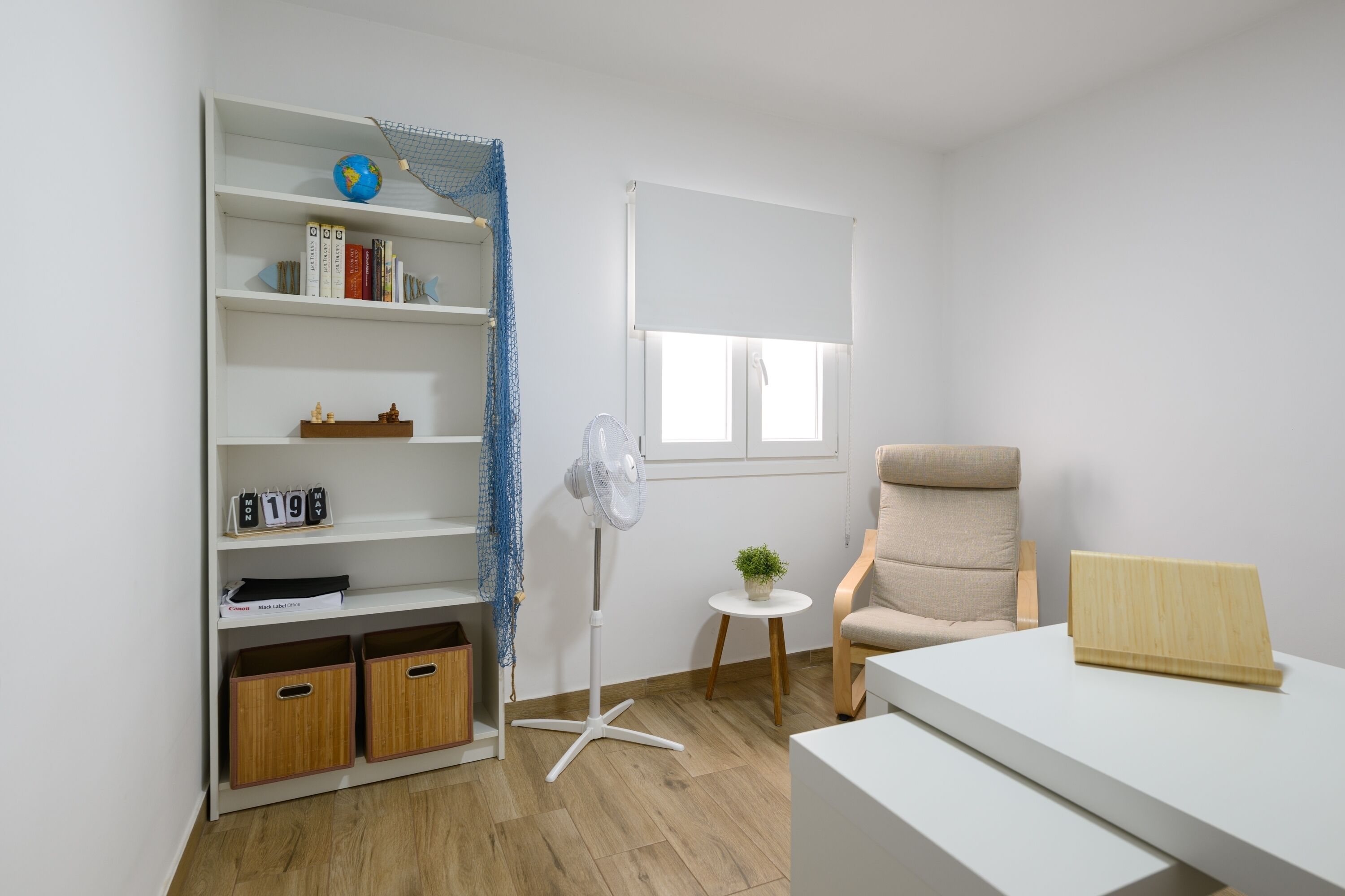 Een kamer met een wit bureau, een boekenkast, een stoel, een kleine tafel met een plant en een staande ventilator.