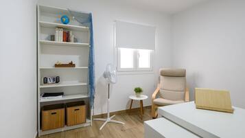Een kamer met een wit bureau, een boekenkast, een stoel, een kleine tafel met een plant en een staande ventilator.