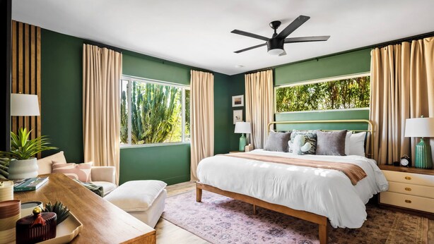 A bedroom with a bed, bedside tables, a desk, a ceiling fan, and large windows with curtains.