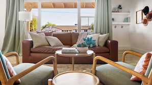 A living room with a brown sofa, two wooden chairs, a round coffee table, and a view of houses through large windows.