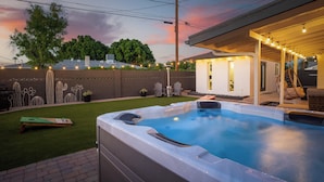 A hot tub in a backyard with a covered patio, a fence with cactus designs, and string lights.