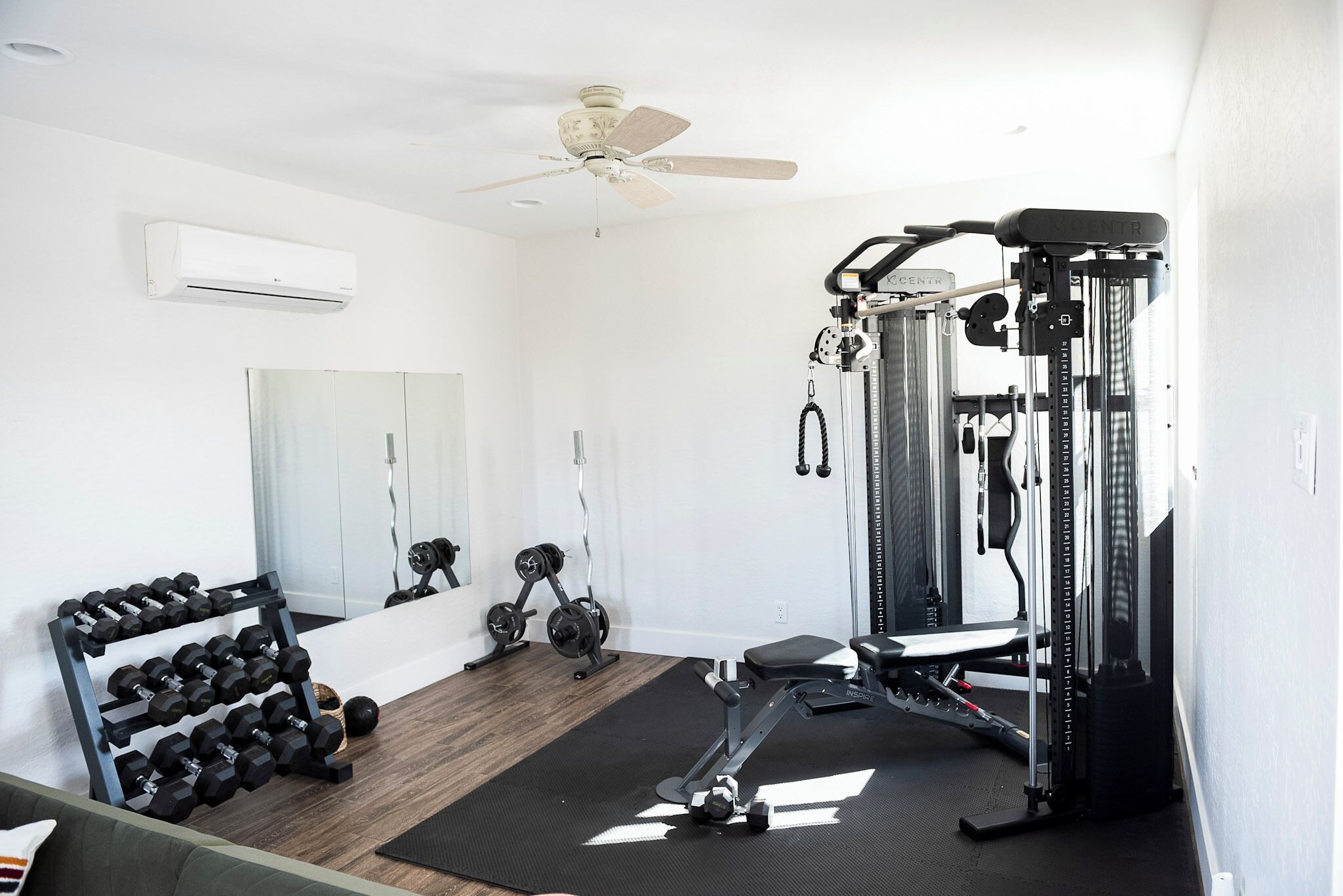 A home gym with a ceiling fan, air conditioning unit, weight rack, dumbbells, and a cable machine.