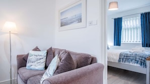 A modern room with a grey sofa, a floor lamp, a bed with a blue and white blanket, and a window with blue curtains.