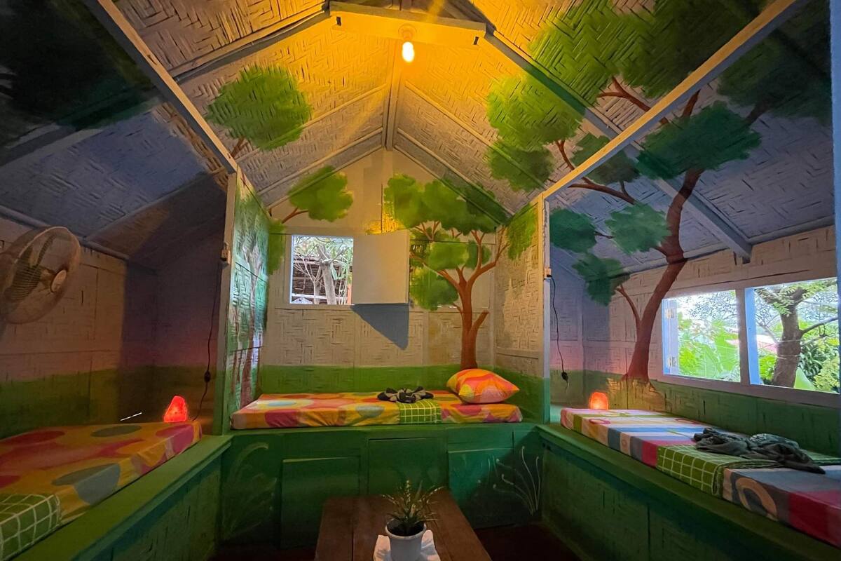 Una habitación con techo inclinado, paredes verdes y una ventana. Hay una cama con ropa de cama colorida y una mesita con una planta.