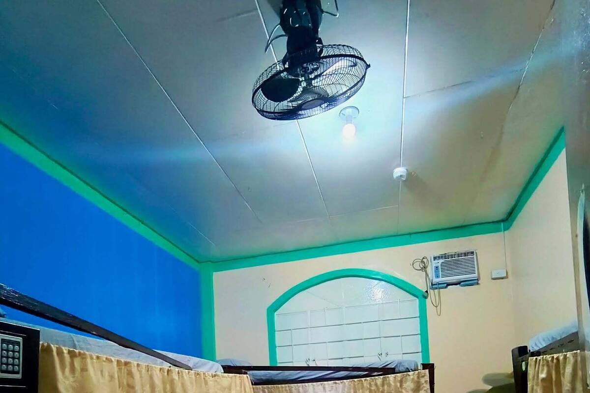 Una habitación con una cama, un ventilador de techo, un ventanal con marco verde y un equipo de aire acondicionado.