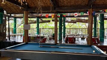 A pool table with a blue felt surface and a Yin-Yang symbol design. The room has a wooden ceiling with hanging light fixtures and green pillars. Flags are strung across the room.