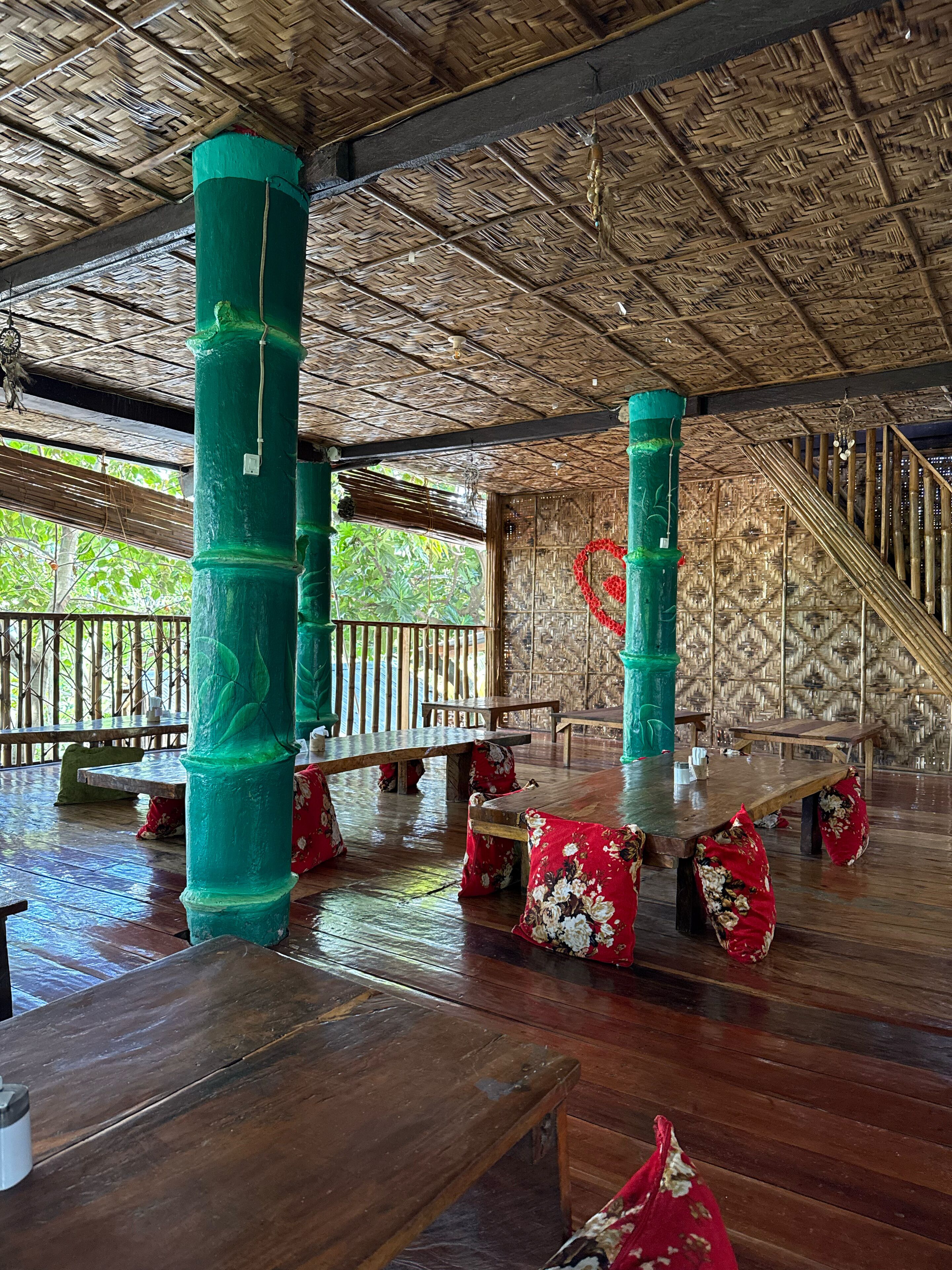An indoor area with wooden tables and benches, green pillars, and a woven ceiling.