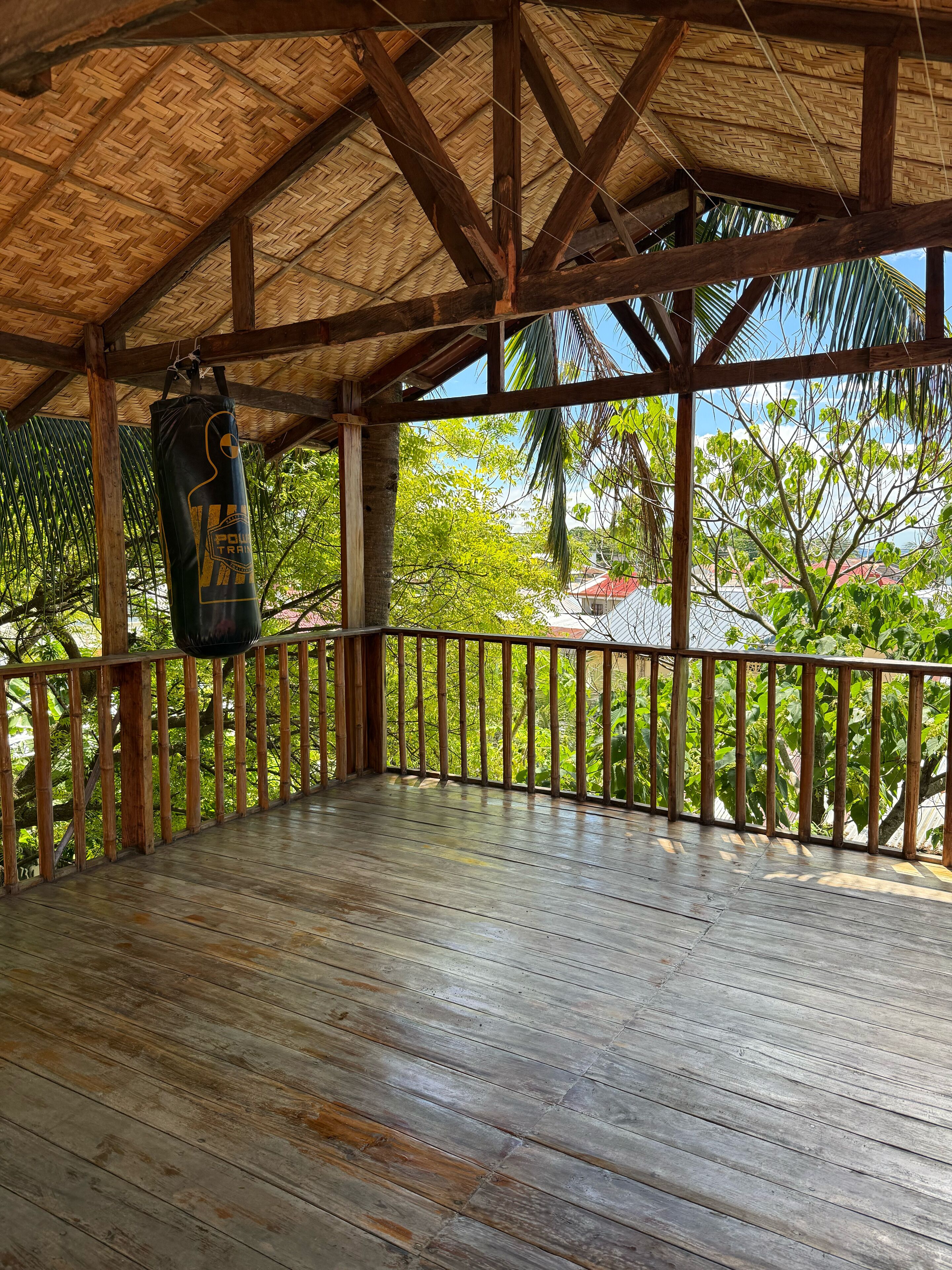 Una terraza de madera con techo de paja, un saco de boxeo y vistas a zonas verdes y edificios.