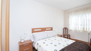 Een slaapkamer met een houten bed, witte beddengoed, een nachtkastje met een lamp, een stoel, en een raam met transparante gordijnen.