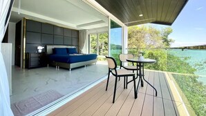 A modern bedroom with a large bed, a bedside table, and a view of a balcony with a table and chairs. - Baan Jayden Duplex Seaview Villa (Phuket)