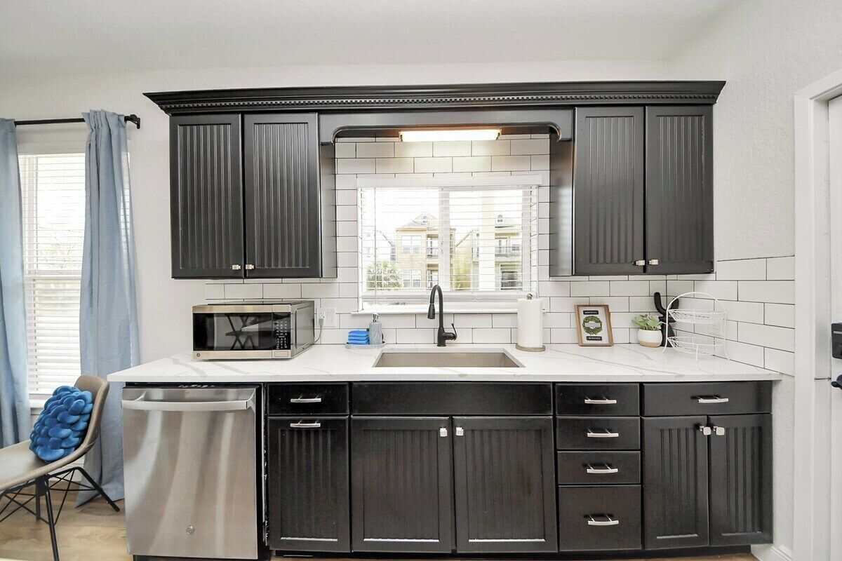 A kitchen with dark cabinets, a white countertop, a microwave, a sink, and a window with a view of buildings.