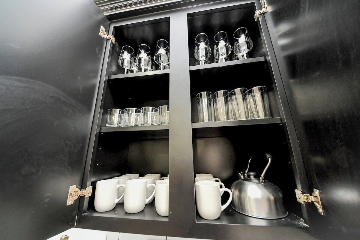 A black cabinet with shelves holding clear glasses, white mugs, and a silver kettle.