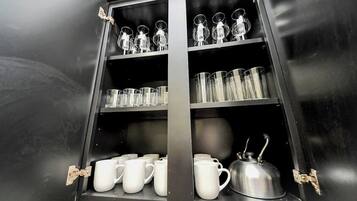 A black cabinet with shelves holding clear glasses, white mugs, and a silver kettle.