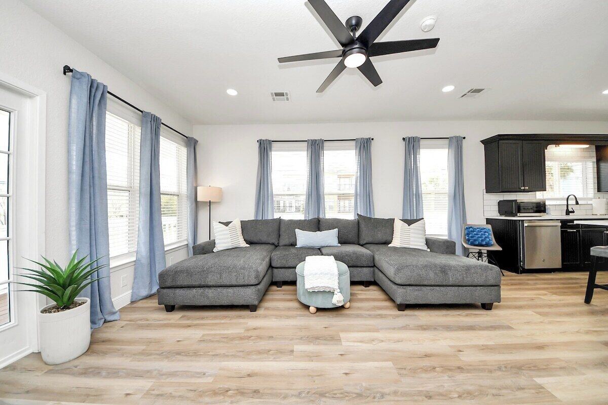 A modern living room with a sectional sofa, a ceiling fan, and a kitchen in the background.