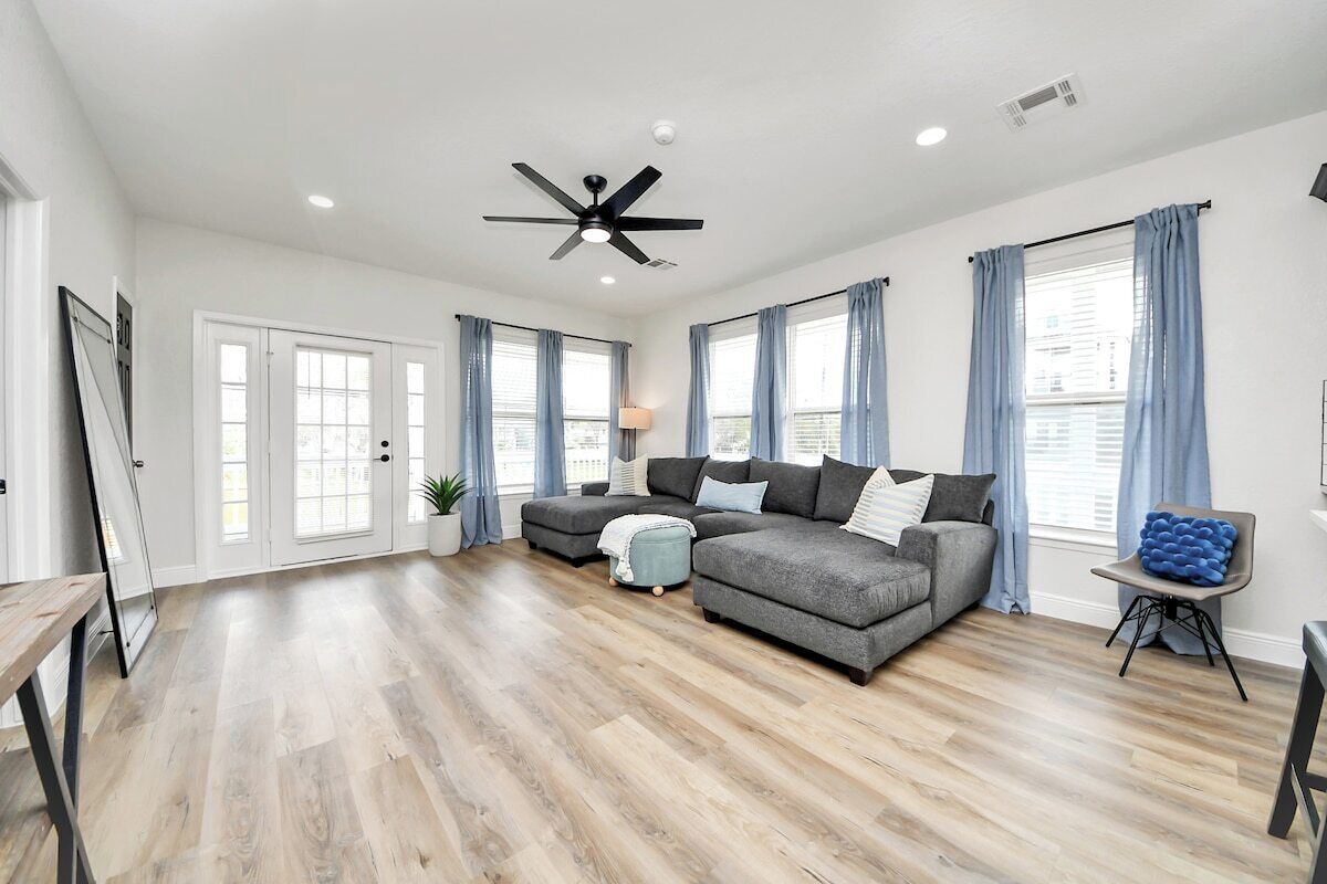 A spacious living room with hardwood flooring, a sectional sofa, a ceiling fan, and large windows with curtains.