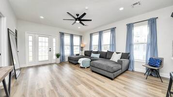 A spacious living room with hardwood flooring, a sectional sofa, a ceiling fan, and large windows with curtains.