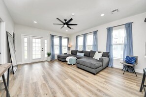 Un salon spacieux doté d’un plancher en bois franc, d’un canapé d’angle, d’un ventilateur de plafond et de grandes fenêtres avec des rideaux.
