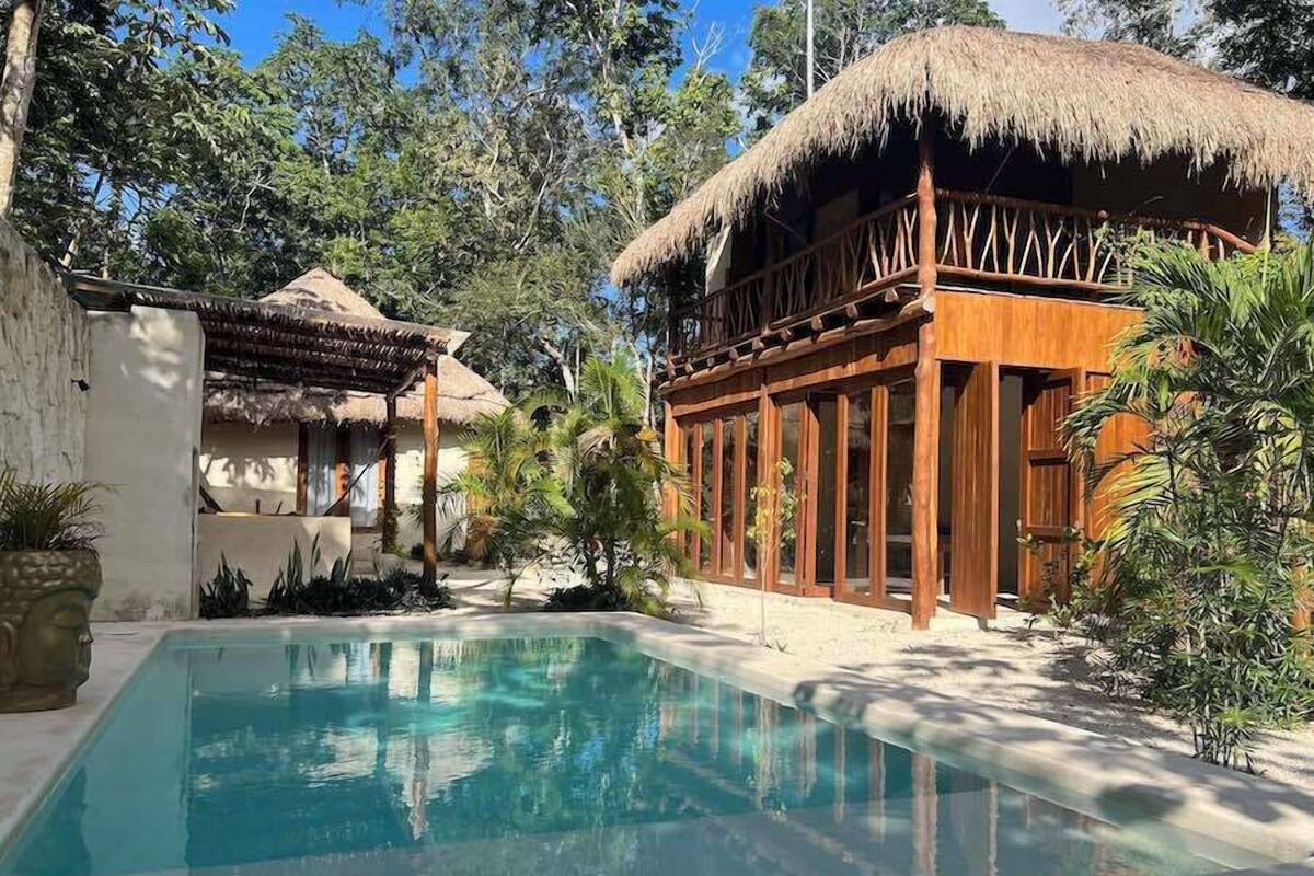Un espace piscine agrémenté d’une structure à toit de chaume, d’un bâtiment en bois doté de grandes fenêtres et d’une végétation luxuriante.