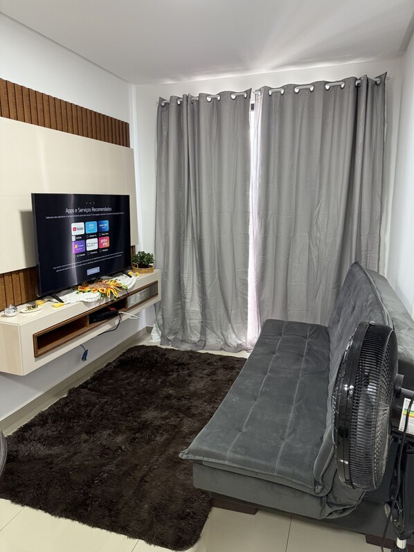 A modern living room with a flat-screen TV mounted on a wooden panel, a grey sofa, a black furry rug, and a standing fan.
