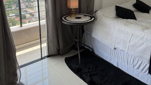 Ein Hotelzimmer mit großem Fenster, einem Bett mit weißer Bettwäsche und schwarzen Kissen, einem kleinen runden Tisch mit Lampe sowie einem flauschigen Teppich.