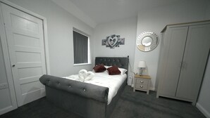 A bedroom with a grey upholstered bed, a bedside table with a lamp, a wardrobe, and a decorative mirror on the wall.