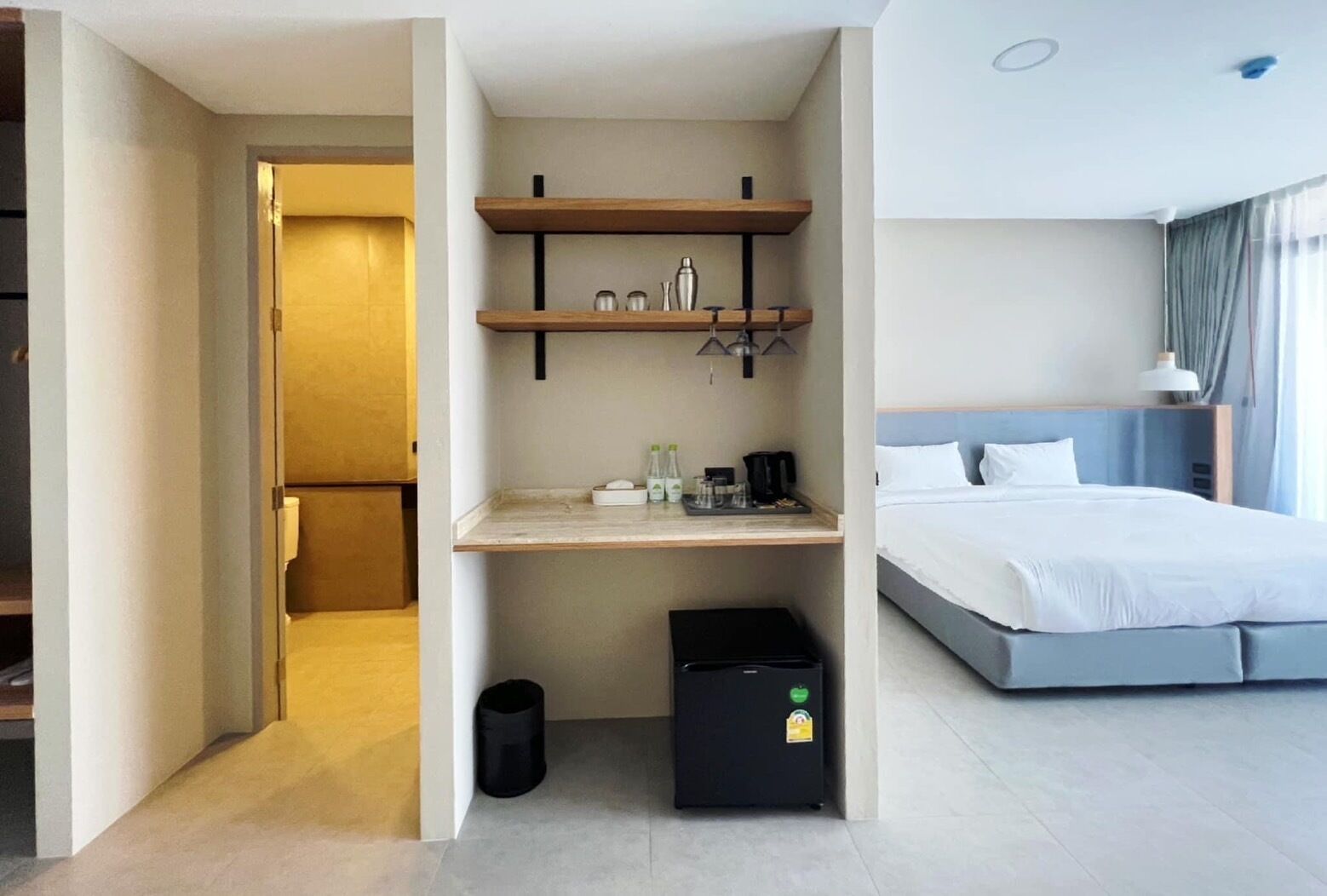 A hotel room with a bed, a desk with a computer, a trash bin, and a small refrigerator.