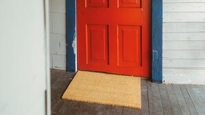 A red door with a doormat in front, wooden floor, and white siding walls. - Cozy Rustic Sailors Room (Houston)