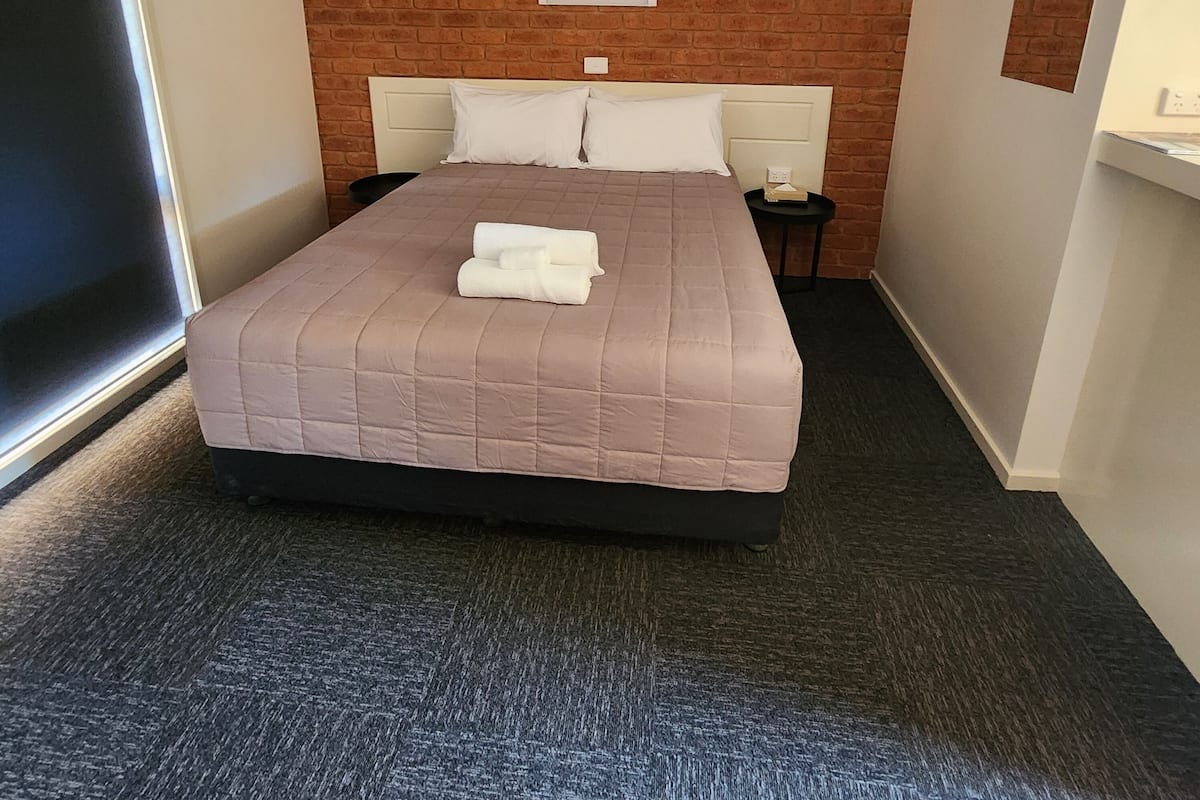 A hotel room with a brick wall, a bed with a beige quilt, two bedside tables, and a window.