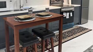 Een moderne keuken met donkergrijze kastjes, een roestvrijstalen koelkast, een magnetron en een fornuis. Er staat een houten tafel met twee zwarte stoelen en een tapijt met een patroon op de vloer.